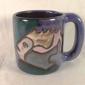 Puede incluir: Una taza de cerámica con un diseño de pez. La taza tiene un asa azul y un cuerpo verde azulado y morado. El pez está delineado en marrón con acentos rosas y blancos.