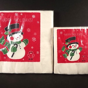 May include: Two packages of holiday napkins. Each napkin features a snowman wearing a black top hat, green scarf and mittens, and holding a cane. The background is red with white snowflakes and the words "HAPPY HOLIDAYS!"