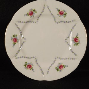 Peut inclure: Une assiette en céramique blanche avec un bord doré et un motif floral. Le motif représente des roses roses et jaunes avec des feuilles violettes.