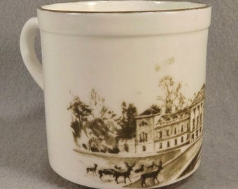 May include: White ceramic mug with a brown illustration of a building and trees. There are deer in the foreground of the image.