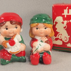May include: A set of two ceramic Christmas salt and pepper shakers. The shakers are shaped like children, one with a red hat and a rocking horse, the other with a green hat and a doll.