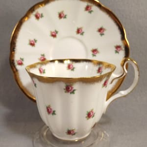 Może przedstawiać: Biały kubek i spodek do herbaty z delikatnym wzorem kwiatowym i złotą obwódką. Kubek i spodek są ozdobione małymi różowymi różami.