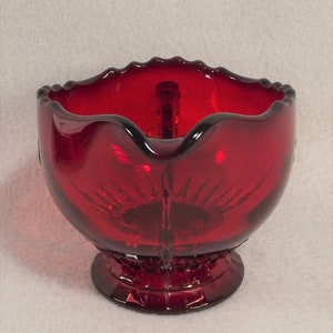 Puede incluir: Una jarra de vidrio roja con un borde festoneado y una base con pie. La jarra tiene un dise&ntilde;o texturizado en forma de sol.