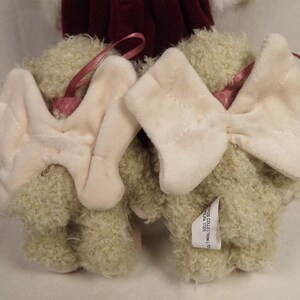 Puede incluir: Dos adornos de osos de peluche con lazos color crema y cintas rosas para colgar. Los osos tienen pelaje verde claro y son parcialmente visibles. Una etiqueta est&aacute; adherida a un oso. El fondo incluye una tela burdeos.