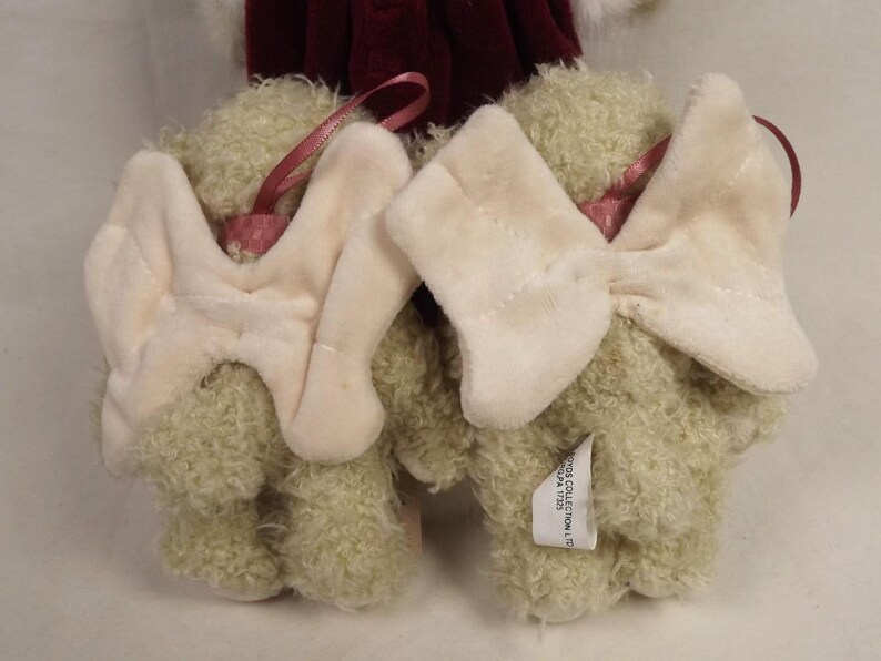 Puede incluir: Dos adornos de osos de peluche con lazos color crema y cintas rosas para colgar. Los osos tienen pelaje verde claro y son parcialmente visibles. Una etiqueta est&aacute; adherida a un oso. El fondo incluye una tela burdeos.