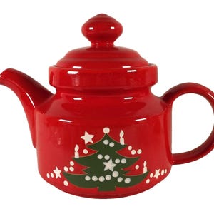 May include: A red ceramic teapot with a lid and handle. The teapot features a green Christmas tree design with white accents and stars. The teapot is a festive item for serving tea during the holiday season.
