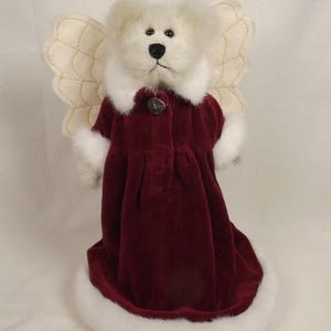 Puede incluir: Una figura de oso de peluche blanco vestida de &aacute;ngel. El oso lleva un vestido de terciopelo burdeos con ribete de piel sint&eacute;tica blanca y un bot&oacute;n decorativo. Tiene alas acolchadas de color crema.