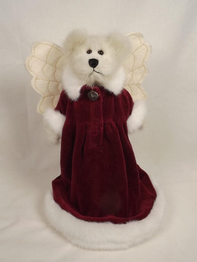 Puede incluir: Una figura de oso de peluche blanco vestida de &aacute;ngel. El oso lleva un vestido de terciopelo burdeos con ribete de piel sint&eacute;tica blanca y un bot&oacute;n decorativo. Tiene alas acolchadas de color crema.