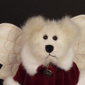 Puede incluir: Un oso de peluche blanco con alas de &aacute;ngel color crema. El oso tiene una nariz negra y ojos marrones. Lleva un abrigo de terciopelo burdeos con un bot&oacute;n decorativo. Las alas est&aacute;n cosidas con hilo dorado.