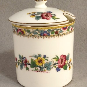 Puede incluir: Un tarro de porcelana blanca con tapa y diseño floral. El tarro tiene un borde dorado y una pequeña cuchara en la tapa. Las flores son en tonos de rosa, amarillo, azul y verde.