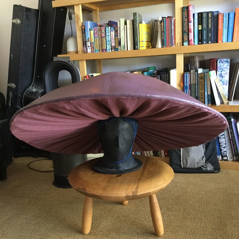 May include: A large, purple mushroom-shaped lampshade with a pleated fabric design. The lampshade is sitting on a wooden stool.