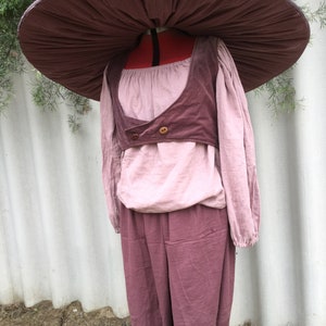 May include: A mauve-colored outfit with a wide, mushroom-shaped hat. The outfit includes a long-sleeved shirt, a vest, and pants. The hat is made of a soft, pliable material.