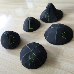 May include: Five black, dome-shaped objects with white markings and green letter labels (A, B, C, D, and E). The objects are arranged on a light-colored wooden surface. The objects appear to be made of a soft material.