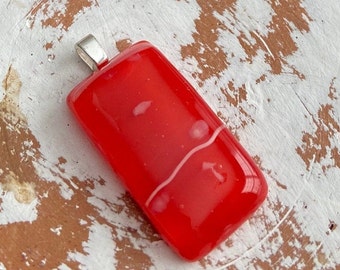 May include: A rectangular red pendant with a silver bail. The pendant has a glossy finish and a white, wavy line design. Small white spots are also visible on the surface. The pendant is likely a piece of jewelry.