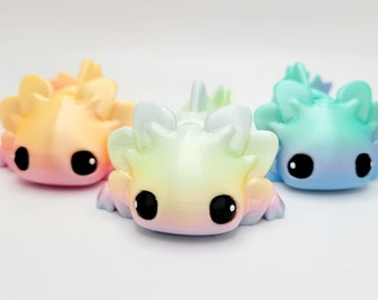 May include: Three small axolotl figurines, each with a different ombre color scheme. The axolotls are made of a smooth, glossy material and have large, black eyes. The colors are a light pink to yellow ombre, a light green to yellow ombre, and a light blue to purple ombre.