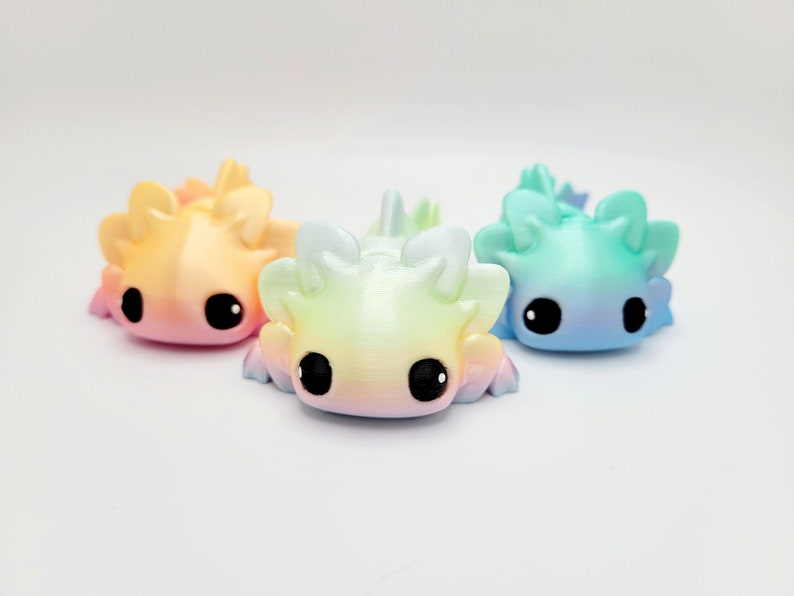 May include: Three small axolotl figurines, each with a different ombre color scheme. The axolotls are made of a smooth, glossy material and have large, black eyes. The colors are a light pink to yellow ombre, a light green to yellow ombre, and a light blue to purple ombre.
