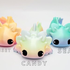May include: Three small, colorful, 3D printed axolotl figurines. The axolotls are in a gradient color scheme. The axolotl on the left is pink and yellow, the middle axolotl is yellow and blue, and the axolotl on the right is blue and green. The axolotls are labeled "Sunset", "Candy", and "Sea".