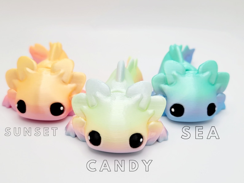 May include: Three small, colorful, 3D printed axolotl figurines. The axolotls are in a gradient color scheme. The axolotl on the left is pink and yellow, the middle axolotl is yellow and blue, and the axolotl on the right is blue and green. The axolotls are labeled "Sunset", "Candy", and "Sea".
