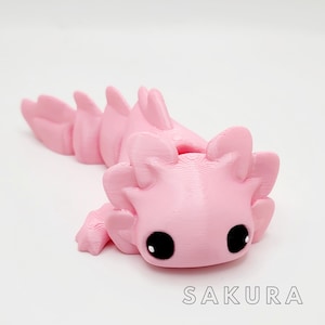May include: A pink 3D printed axolotl figurine with black eyes. The figurine is lying on its belly with its tail curled up.