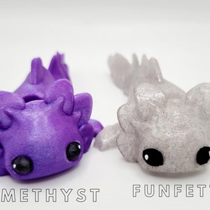 May include: Two 3D printed axolotl figurines, one purple with glitter and the other white with glitter. The purple axolotl is labeled "Amethyst" and the white axolotl is labeled "Funfetti".