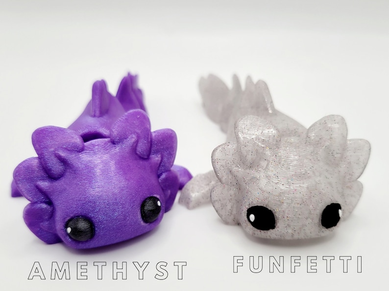 May include: Two 3D printed axolotl figurines, one purple with glitter and the other white with glitter. The purple axolotl is labeled "Amethyst" and the white axolotl is labeled "Funfetti".