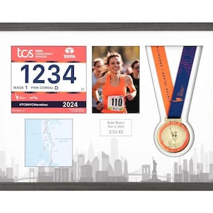 May include: Framed New York City Marathon memorabilia. Includes a race bib with the number 1234, a photo of a runner wearing number 110, a medal, and a course map. The frame has a gray finish and a cityscape silhouette.