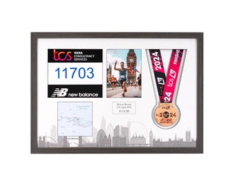 May include: A framed display with a runner's medal, bib number, and photo. The medal is bronze with a pink and black ribbon. The bib number is 11703. The photo shows a runner crossing the finish line. The frame has a gray border and a white background. The text on the medal reads "tcs London 2024" and "24 tcs 27 FINISH".