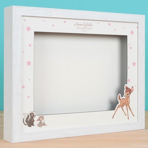 Puede incluir: Marco de fotos de madera blanco con un paspartú rosa y blanco. El marco tiene un recorte para una foto y presenta un diseño de estrellas rosas. El marco también tiene un recorte de Bambi,  Tambor y Flor de la película de Disney Bambi. El texto "Emma White" está impreso en el marco.