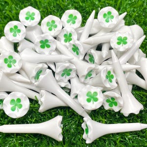 May include: A set of white golf tees with green four-leaf clover designs. The tees are arranged in a pile on a green grassy surface.