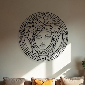 May include: A black metal Medusa head wall art piece, mounted above a beige sofa with orange and beige cushions. The room is decorated with plants, a wooden coffee table, and a neutral-coloured rug.