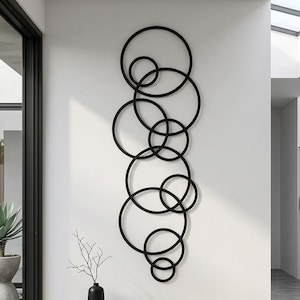 May include: A vertical wall art piece consisting of multiple interconnected black metal rings of varying sizes arranged in a cascading, overlapping pattern. The sculpture is mounted on a plain white wall above a low wooden bench in a modern interior.