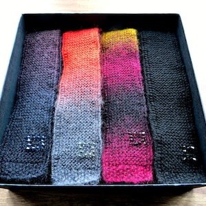 May include: Four knitted headbands in a black box. The headbands are ombre, with colors transitioning from black to gray, gray to red, red to pink, and pink to black.