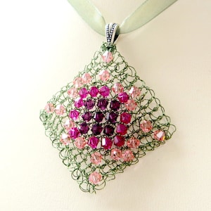 May include: A green crocheted pendant with pink and purple crystals. The pendant is attached to a green ribbon.