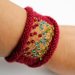 K&ouml;nnte beinhalten: Ein gestricktes bordeauxfarbenes Armband mit einem goldfarbenen Drahtgeflecht-Mittelst&uuml;ck. Das Geflecht ist mit farbigen Perlen in Rot-, Blau- und Gr&uuml;nt&ouml;nen verziert. Das Armband ist im Manschettenstil gehalten und wird am Handgelenk getragen.