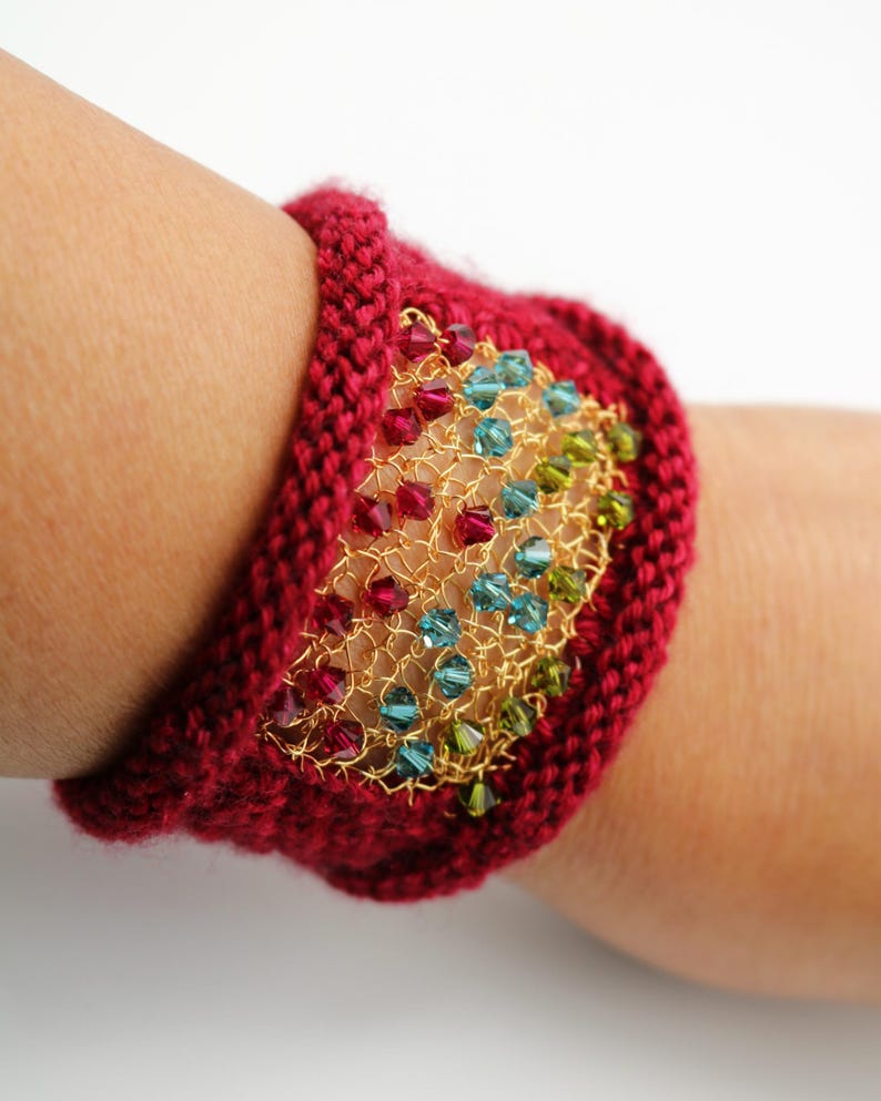 K&ouml;nnte beinhalten: Ein gestricktes bordeauxfarbenes Armband mit einem goldfarbenen Drahtgeflecht-Mittelst&uuml;ck. Das Geflecht ist mit farbigen Perlen in Rot-, Blau- und Gr&uuml;nt&ouml;nen verziert. Das Armband ist im Manschettenstil gehalten und wird am Handgelenk getragen.