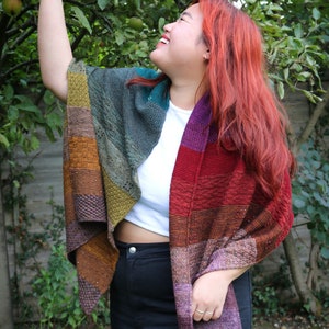 May include: A woman wearing a colourful striped knitted shawl with a white shirt and black trousers. She is reaching up to touch an apple on a tree.