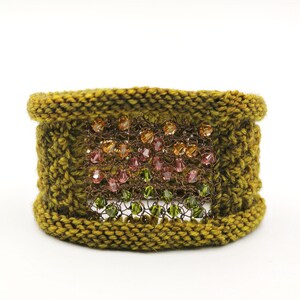 K&ouml;nnte beinhalten: Ein handgefertigtes olivgr&uuml;nes Strickarmband. Das Armband hat ein zentrales Feld aus verflochtenem Metall mit bunten Perlen in Orange, Rosa und Gr&uuml;n. Ein einzigartiges Accessoire.