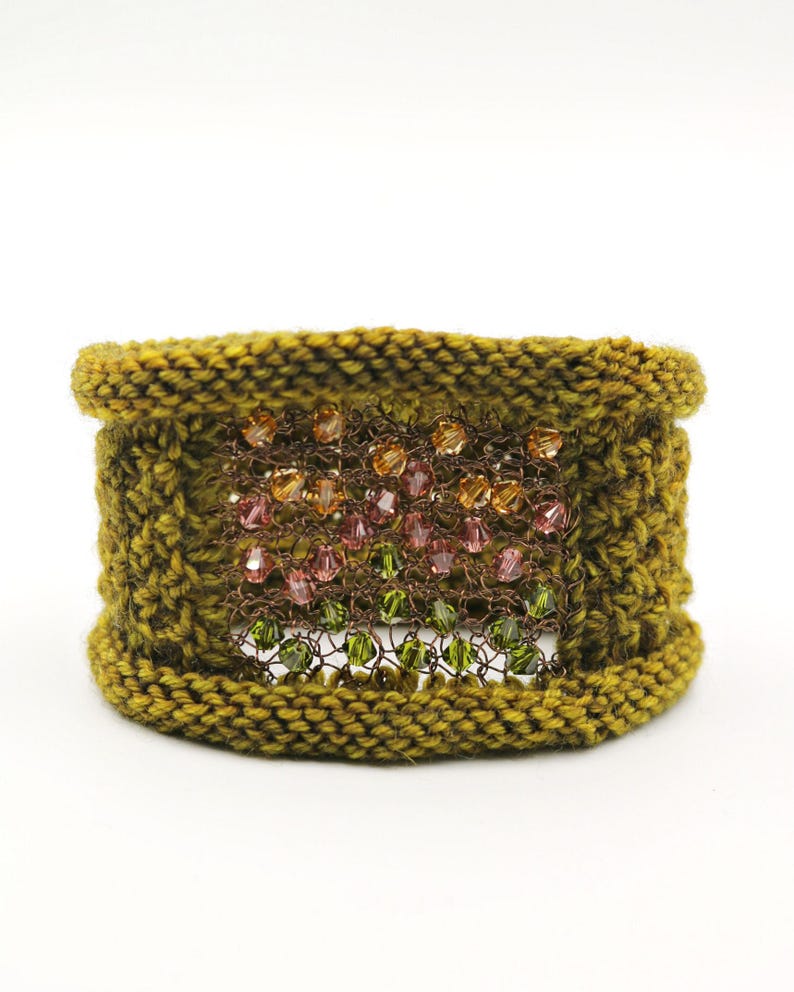 K&ouml;nnte beinhalten: Ein handgefertigtes olivgr&uuml;nes Strickarmband. Das Armband hat ein zentrales Feld aus verflochtenem Metall mit bunten Perlen in Orange, Rosa und Gr&uuml;n. Ein einzigartiges Accessoire.