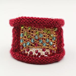 K&ouml;nnte beinhalten: Ein gestricktes Armband in tiefroter Farbe. Das Armband hat ein goldfarbenes Mesh-Panel, das mit farbigen Perlen in Gr&uuml;n, Blau und Rot verziert ist. Das Armband ist ein breites Band, geeignet f&uuml;r ein Handgelenk. Das Design ist handgefertigt.