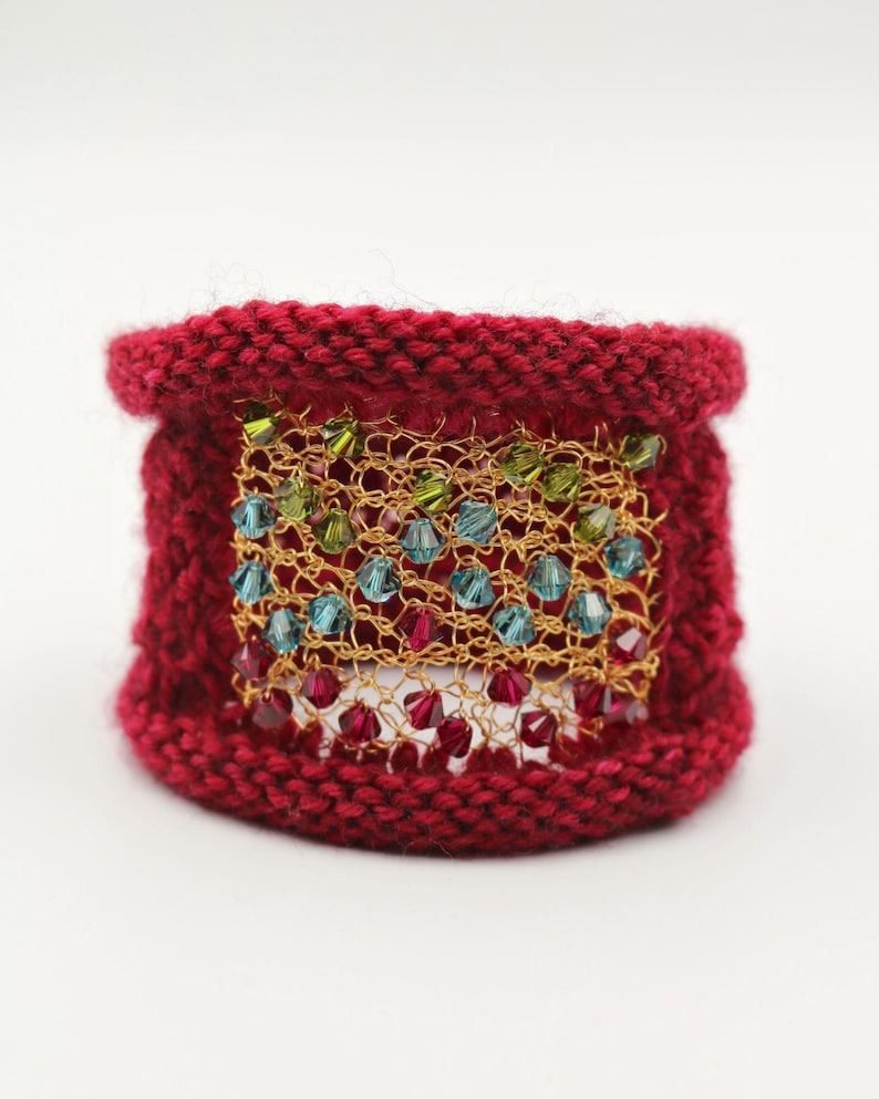 K&ouml;nnte beinhalten: Ein gestricktes Armband in tiefroter Farbe. Das Armband hat ein goldfarbenes Mesh-Panel, das mit farbigen Perlen in Gr&uuml;n, Blau und Rot verziert ist. Das Armband ist ein breites Band, geeignet f&uuml;r ein Handgelenk. Das Design ist handgefertigt.