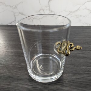 May include: A clear glass tumbler with a gold-colored snake figurine on the rim.