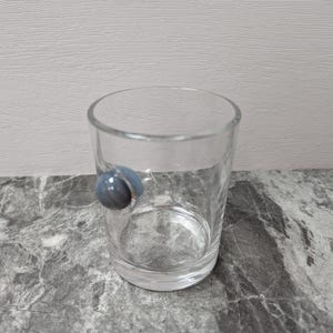 May include: A clear glass tumbler with a blue marble-like accent on the side. The glass is cylindrical and appears empty, set against a grey and white marbled surface.
