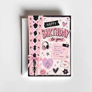May include: A pink and black birthday card with the words "HAPPY BIRTHDAY to you!" in a playful font. The card features a corset-style design with pink ribbons and a bow, along with various illustrations like hearts, stars, and a smiley face.