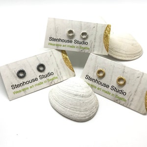 May include: Three pairs of small, simple, minimalist earrings on white cards. The cards have the text "Stenhouse Studio" and "Wearable art made in Seattle". The earrings are silver, gold, and black.