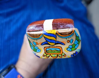 May include: A hand-painted wooden object with a brown top and a colorful face design. The face features blue, yellow, orange, and green accents. A white stripe wraps around the center of the object.