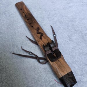 May include: A wooden Native American style flute featuring a light brown wood grain body with a dark brown mouthpiece and block. The instrument includes a carved snake design, leather lacing with silver-toned beads, and six finger holes along the top.