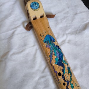 May include: A light-coloured wooden flute featuring a hand-painted blue and purple jellyfish design with flowing tentacles. The instrument includes a block with a circular water symbol, a dark mouthpiece, and brown leather ties near the top.