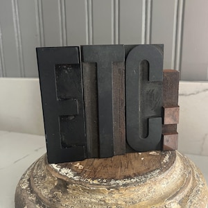 May include: Three individual black letterpress blocks arranged to spell the letters E, T, and C, featuring a matte finish. The blocks are positioned upright on a circular, weathered wooden base with a visible grain and a distressed, light-colored texture.