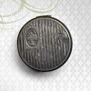 May include: A round, silver-toned metal compact case featuring a vertical striped pattern. The lid displays an embossed oval frame containing a floral basket design and the text "CARA NOME" printed in a rectangular block on the right side.