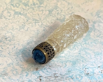 May include: A clear glass perfume bottle with a textured floral pattern. The bottle has a brass-colored metal collar and a blue, faceted glass stopper. The bottle is lying on a light blue patterned surface.